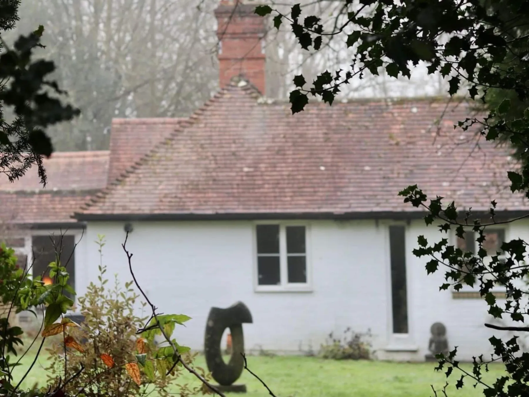 The white studio with its tiled roof and brick chimney seen across the lawn through holly branches on a misty morning.