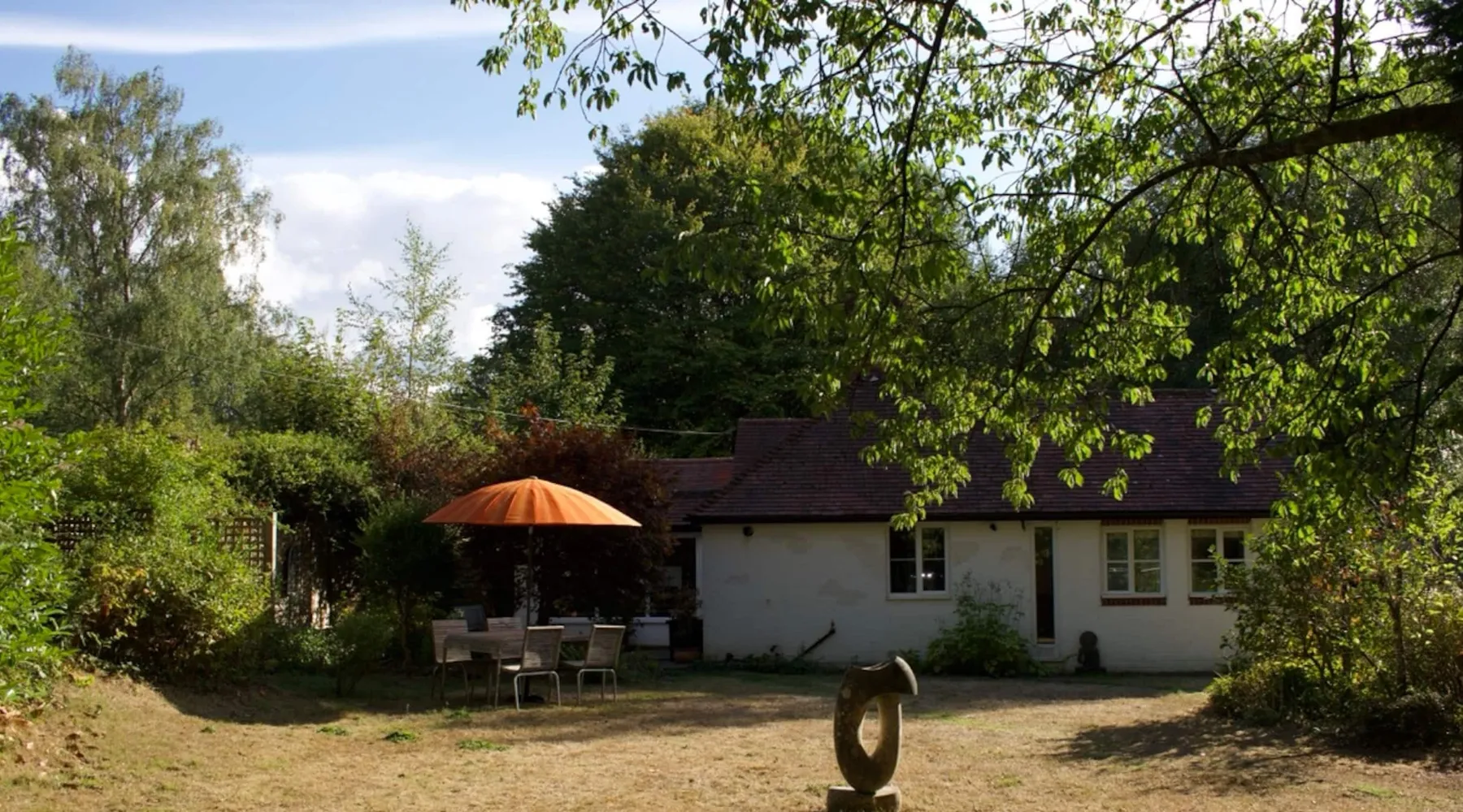 The white studio across the front lawn on a summer afternoon, with a stone sculpture on the grass and a table under an orange parasol.